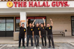 Five men in black shirts with arms stretched above their heads, standing in front of a Halal Guys restaurant