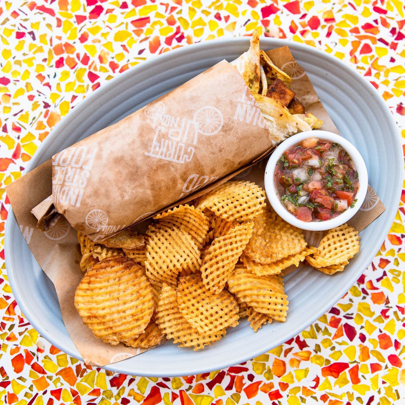 indian food franchise wrap and fries with chips on plate sitting onto of colorful tabletop