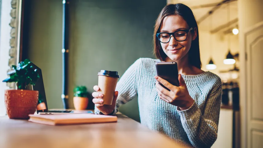 Young lady in glasses holding a coffee looking at her cell phone