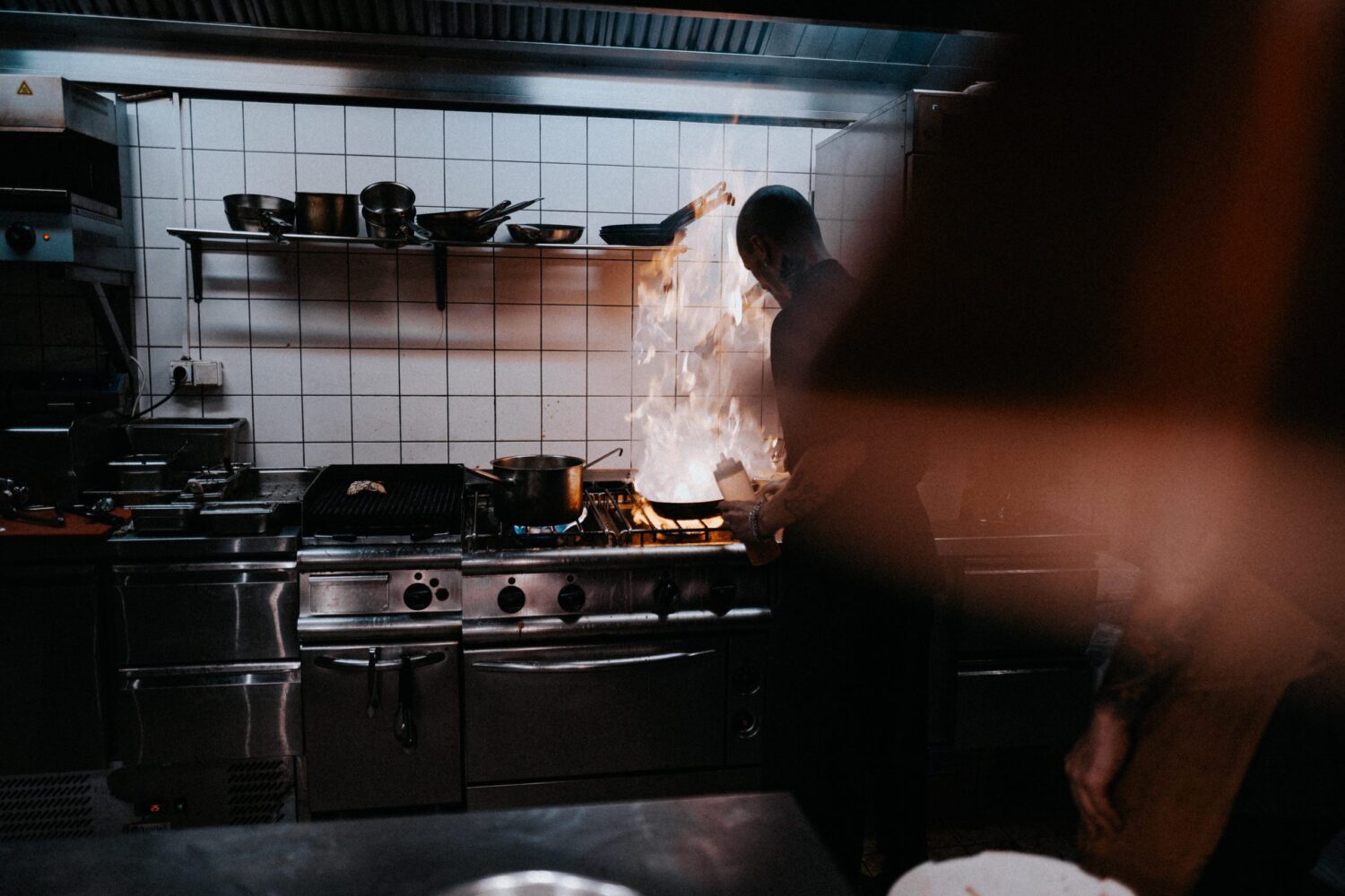 A dark image of man standing at grill, large flame coming from stovetop pan